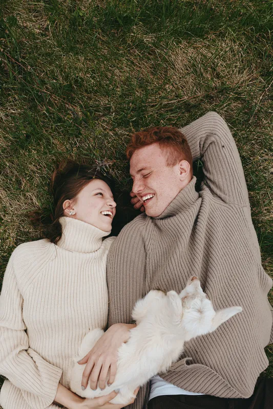 Couple lies on grass wearing cozy sweaters, sharing smiles with a playful goat kid between them, creating a warm intimate lifestyle scene outdoors on a sunny day.