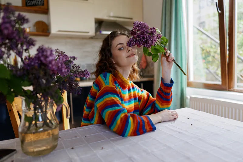Woman in a colorful striped sweater sits at a kitchen table, holding a bouquet of purple flowers near a window, conveying a warm home lifestyle and cheerful intimate moment