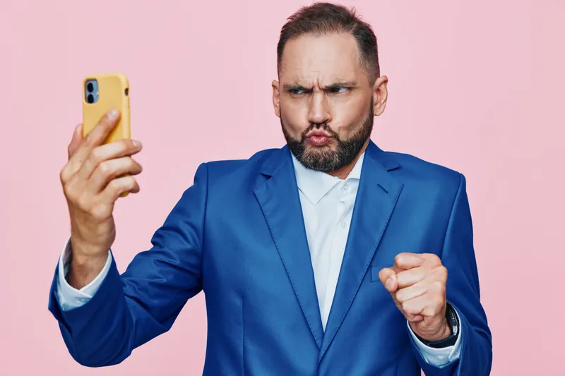 A professional man in a blue suit poses for a selfie against a solid pink background, showcasing business focus and a confident, assertive mood.