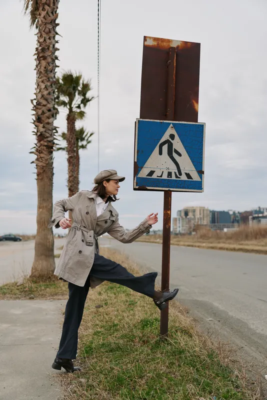 Person in beige coat and hat playfully kicking a pedestrian crossing sign outdoors near palm trees on roadside. Concept of urban life, humor, and casual fashion style in nature.