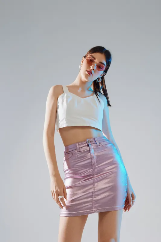 Young woman beauty fashion model wearing white crop top and shiny pink skirt poses confidently against a neutral background. Stylish sunglasses and earrings complete her look.