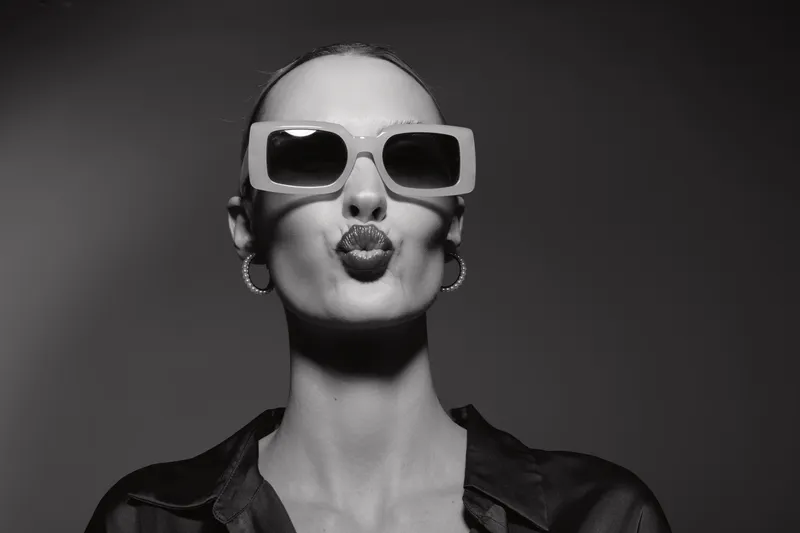 Stylish white woman with pale skin wears large sunglasses and hoop earrings, making a kiss face in black shirt. Concept for fashion, glamour, and confident attitude.