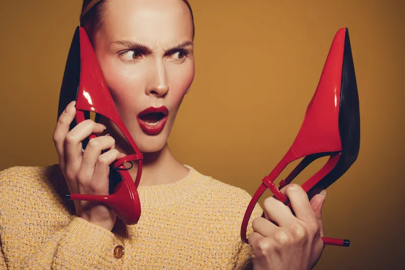Surprised woman in yellow sweater holds red high heel shoes near face, expressing shock and confusion. Studio portrait with warm background and dramatic facial expression concept.