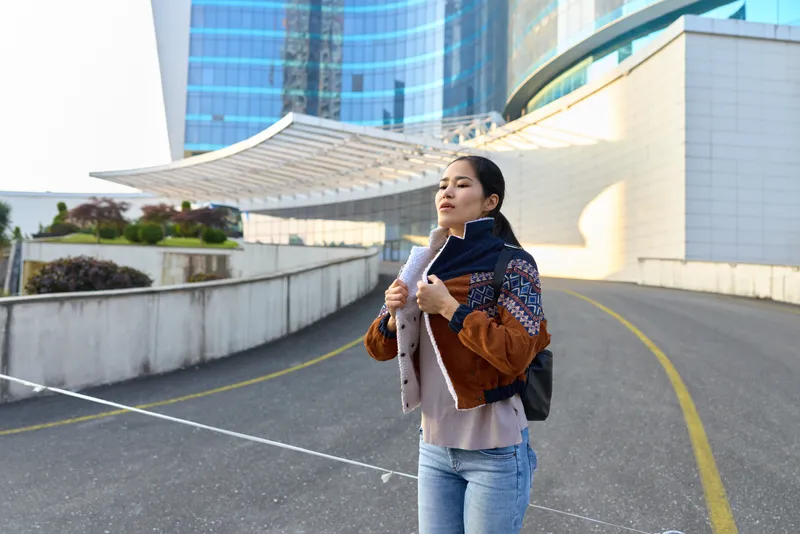 Female city traveler wearing a warm jacket scarf and backpack walking along a modern urban avenue with glass towers in view casual streetwear moment and confident stride