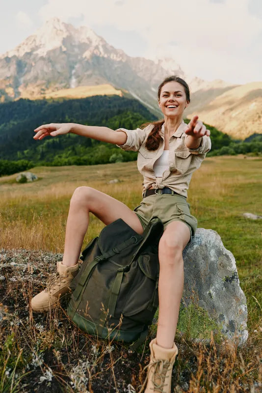 Cheerful woman hiking outdoors with backpack sits on a rock in mountain landscape, casual outfit, enjoying sunny day and freedom, lifestyle portrait