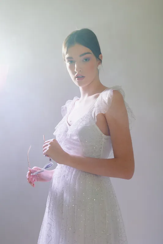 Young woman in a white lace dress holding sunglasses looks thoughtfully at the camera. Soft lighting highlights her natural beauty and fashion style. Studio portrait concept.