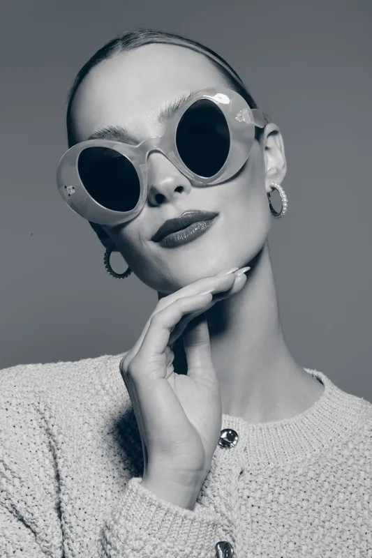 Stylish white woman with pale skin wearing large round sunglasses and earrings poses with hand near chin on gray background.