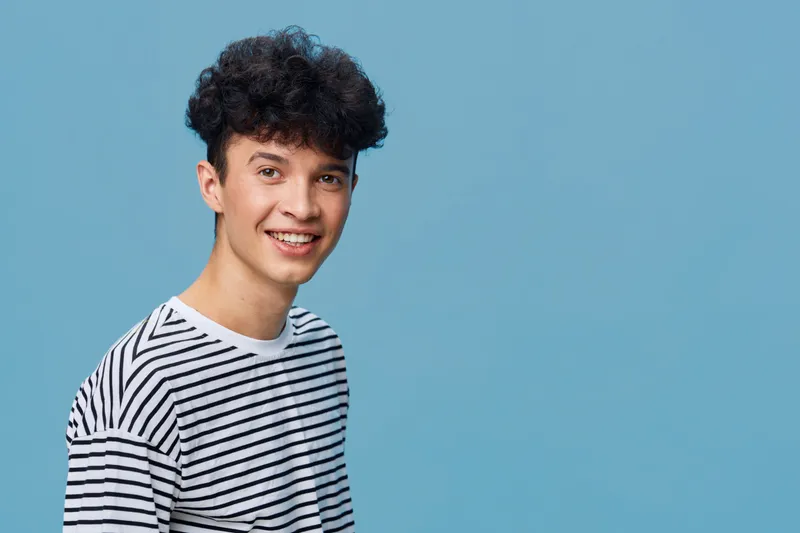 smiling young man with curly hair wearing striped shirt on blue background expressing happiness and friendliness in casual style