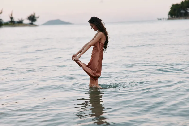 Woman standing in shallow sea water wearing a flowing dress, enjoying peaceful sunset with calm mood and natural beauty.