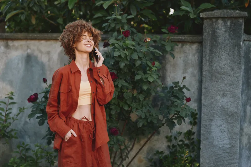 Cheerful woman outdoors, smiling on the phone, warm sunlight, casual orange outfit, relaxed pose, garden backdrop, natural colors, lifestyle moment in warm light