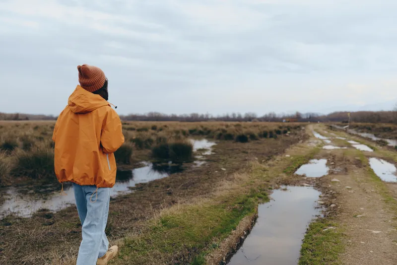 woman lifestyle outdoors scene, hooded jacket and jeans, walking on muddy path beside river, nature landscape with cloudy sky, winter fields and puddles, calm back view