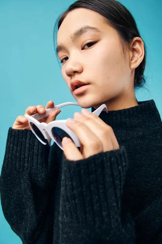 Young woman with dark hair, wearing a black sweater, holding white sunglasses near her face against blue background. Closeup portrait of a trendy female model with subtle makeup and natural light.