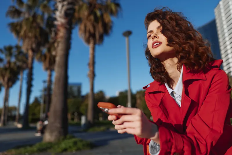 Young woman with curly hair enjoys sunny weather while holding a sunglasses case outdoors near palm trees with a relaxed and happy expression.
