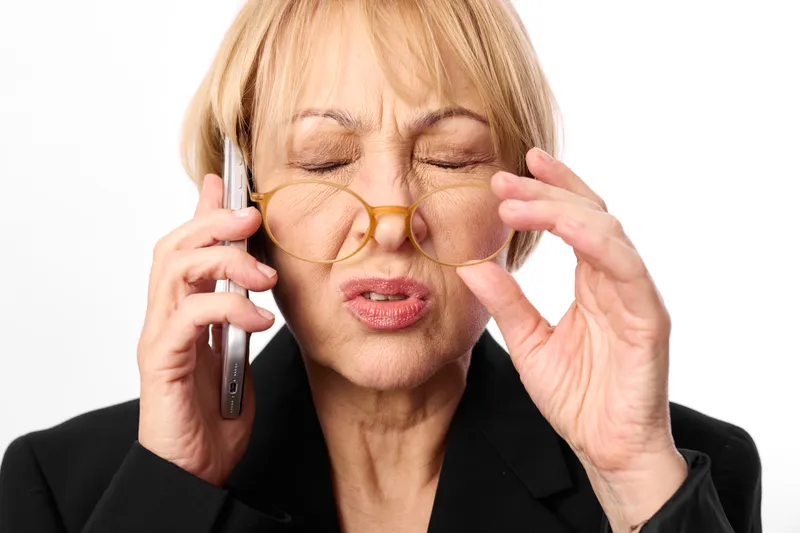 mature woman with blonde hair wearing eyeglasses, holding a phone to her ear, eyes closed, thoughtful expression, studio white background, business communication, calm mood