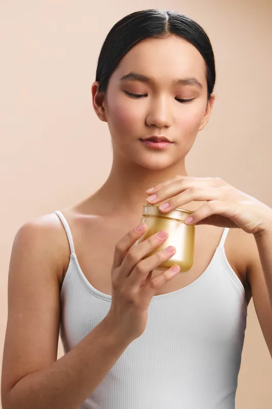 asian woman holds a small jar of cream in a calm studio, focusing on skincare with a gentle expression, soft light, clean setting, and a sense of wellness and self care.