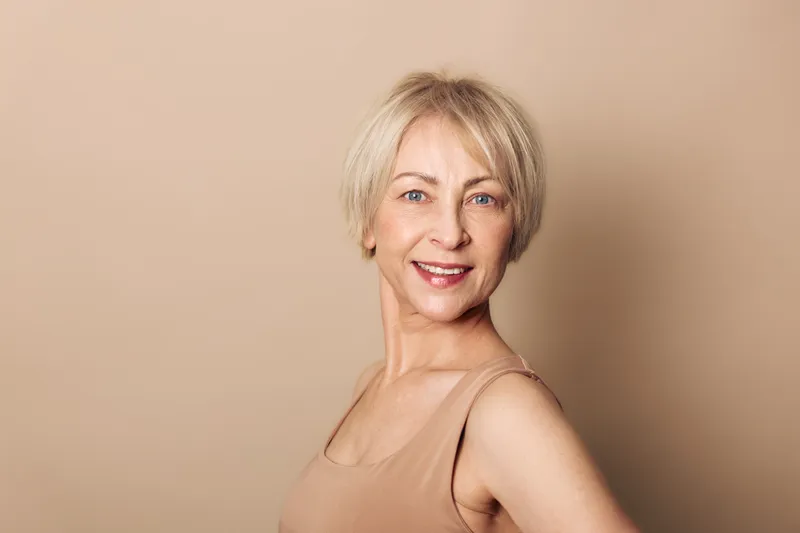 Smiling mature woman with short blonde hair wearing beige top poses confidently against neutral background. Concept of natural beauty, aging gracefully, wellness, and self-confidence in studio.