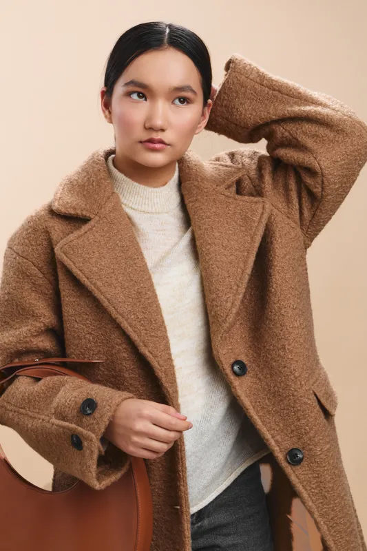 Asian woman wearing a warm brown coat stands poised in a soft studio setting, conveying style and confidence. Her calm expression and chic outfit create a modern, sophisticated mood for fashion and