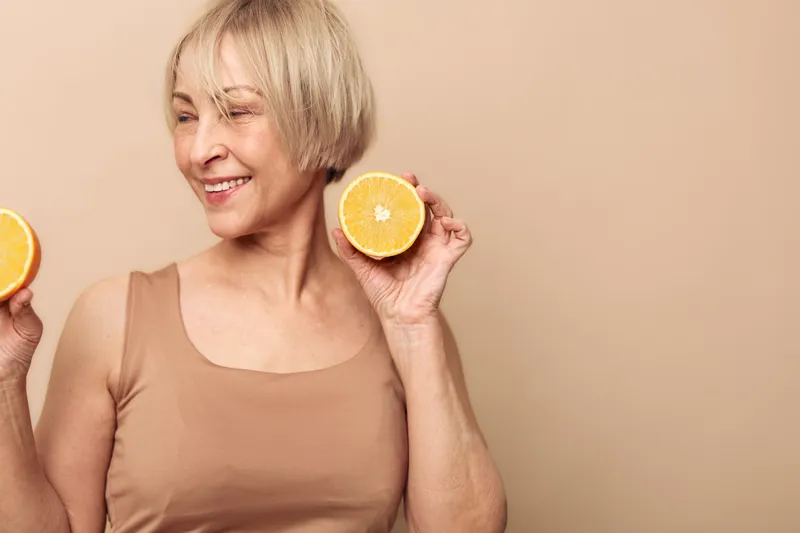 Smiling mature woman with short blonde hair holds two orange halves near face on beige background. Concept of healthy lifestyle, natural vitamin C, wellness and joyful aging.