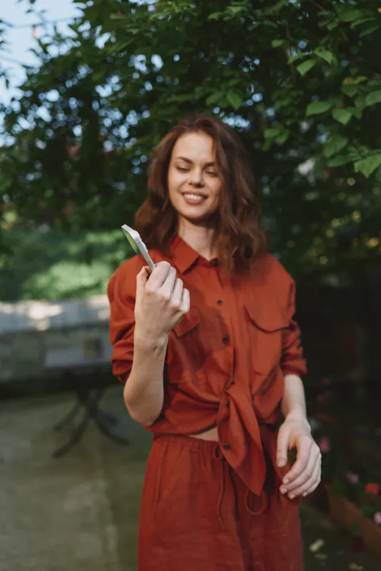 Young woman in an orange dress holds a small brush outdoors, smiling in a sunlit garden, standing among green trees in a candid moment of self care and creativity