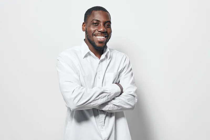 African American man with dark skin wearing white shirt smiling with arms crossed standing against white background. Confident male portrait with casual style and positive expression.