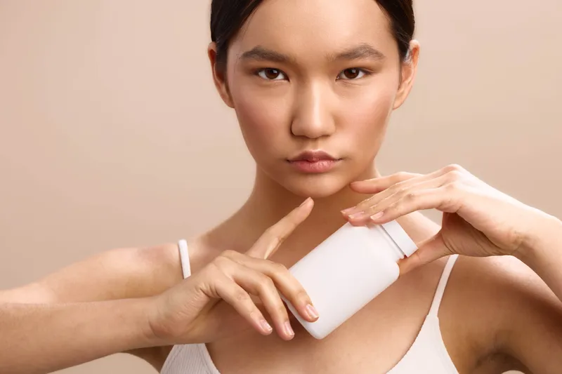 Asian woman applies lotion from a bottle on her skin, showcasing a quiet skincare routine in a clean, minimal setting, conveying calm confidence and personal wellness through a graceful, focused
