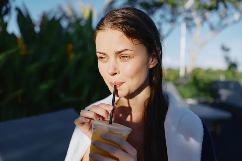 Young woman with wet hair drinks orange juice through a straw outdoors in bright sunlight wearing a white robe with a relaxed lifestyle mood in natural summer environment.