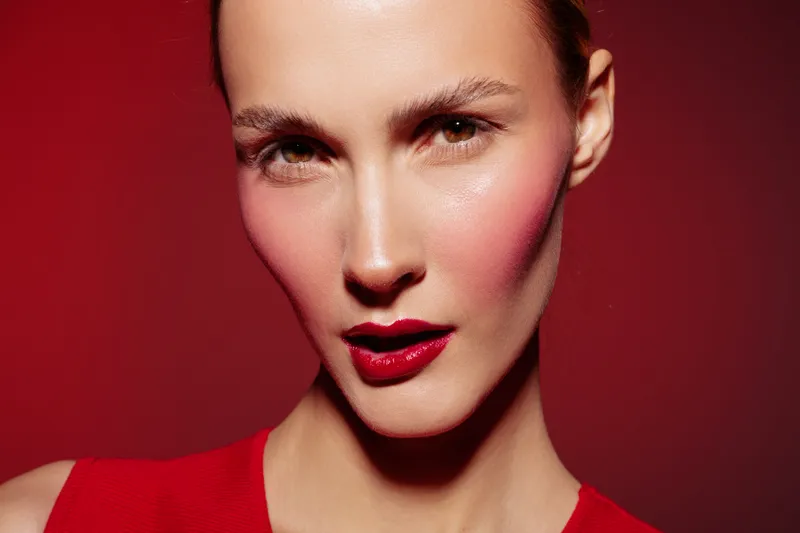 Close-up of a woman with red lipstick and blush on her cheeks wearing a red top against a red background. Concept of beauty, makeup, fashion, and confident expression.