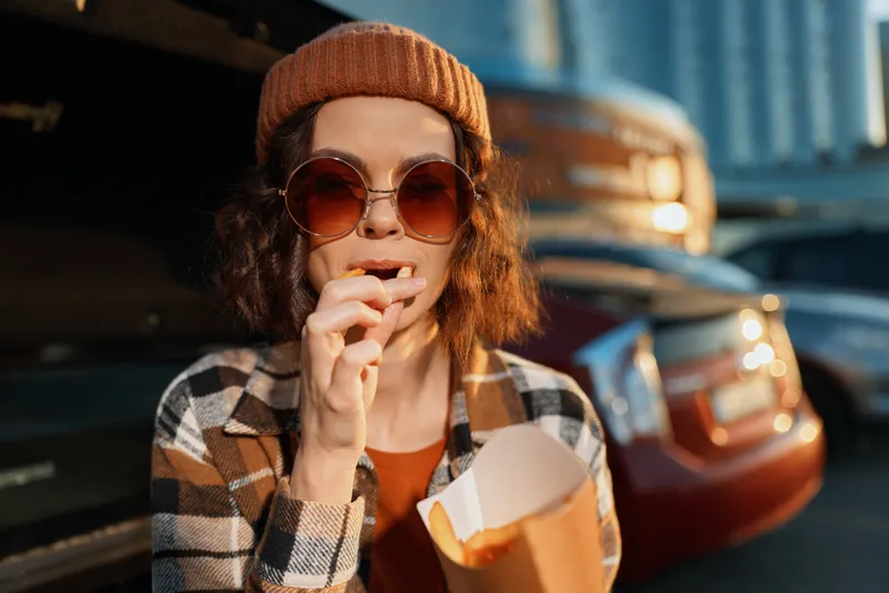 Woman eats fries from paper container by car trunk, wearing sunglasses and beanie in urban goldenhour light, candid lifestyle portrait with authenticity and emotional storytelling.