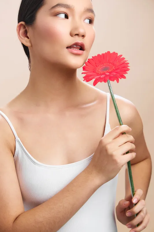 Asian woman gently smiles as she holds a bright pink flower near her face, creating a calm, fresh mood in a simple studio setting suitable for beauty and lifestyle campaigns.