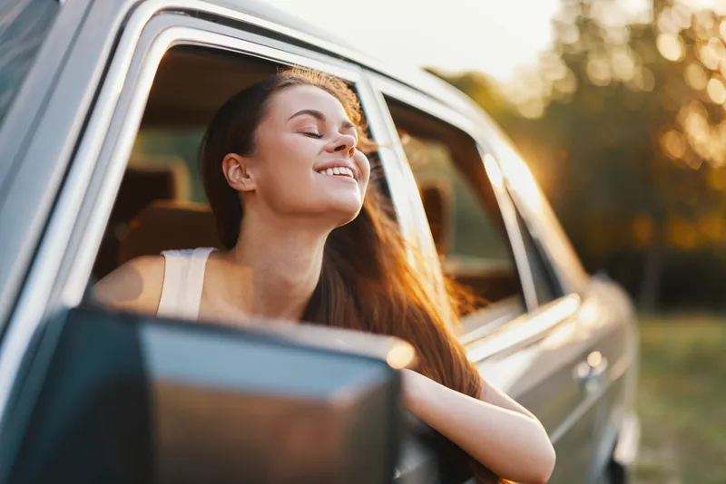 Happy young woman enjoying freedom and fresh air from a vintage car, with sunlit trees in the background, representing travel, joy, and adventure.