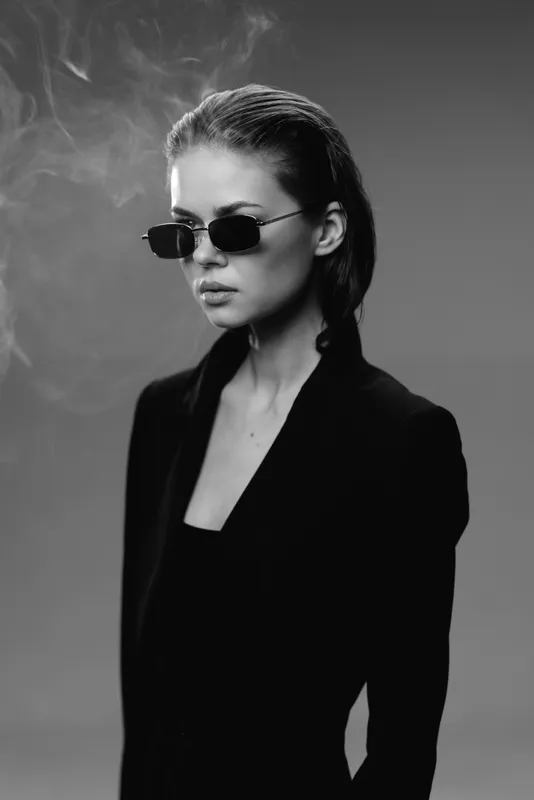 Stylish woman wearing sunglasses and black blazer in moody black and white portrait with smoke effects and sleek hair.
