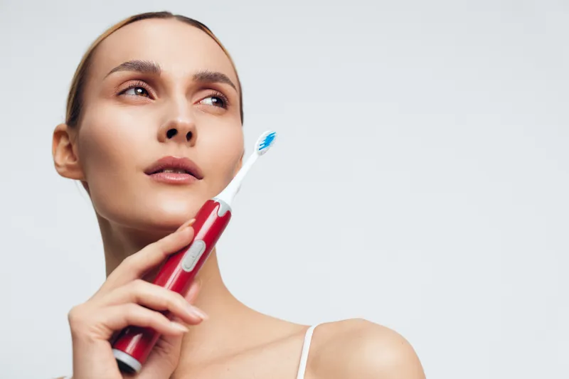 Close-up of a woman with clear skin holding a red electric toothbrush near her face, illustrating oral care, hygiene, and daily beauty routine. Soft light creates a calm mood.