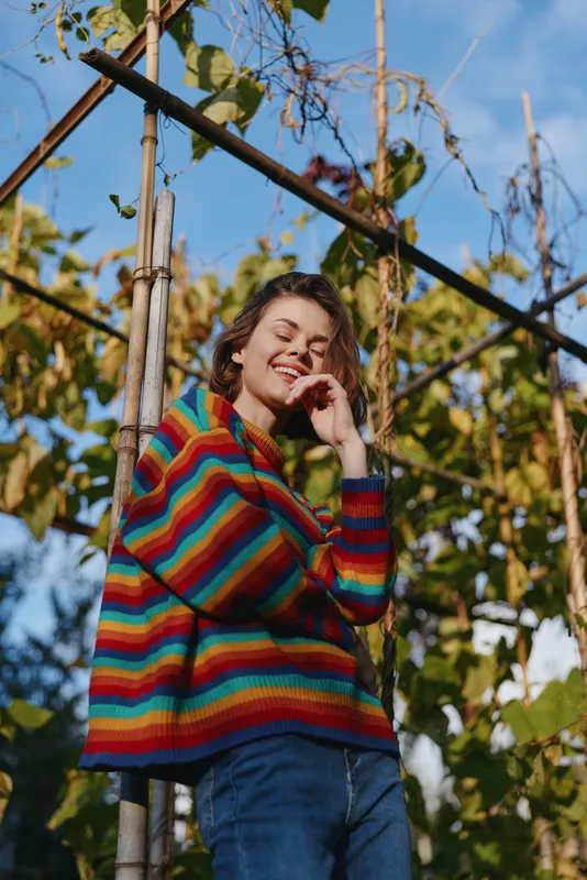 Young woman in her early 20s wearing a colorful striped sweater, smiling and leaning on a wooden trellis in a sunny garden with blue sky, casual outfit and relaxed lifestyle vibe.