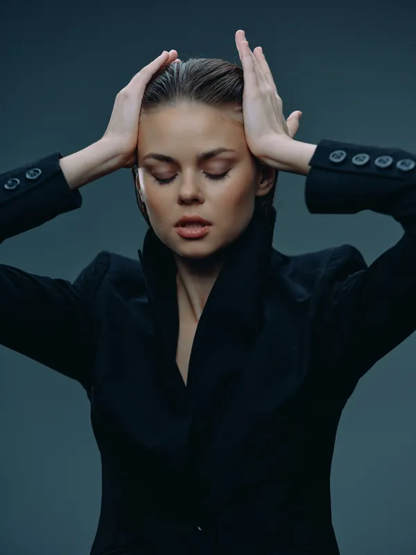 Young woman in black blazer with closed eyes holding her head showing stress and fatigue.