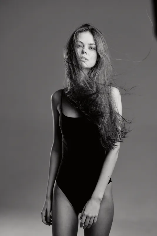 Black and white portrait of a slender woman with long hair wearing a black bodysuit in a minimalist studio setting.