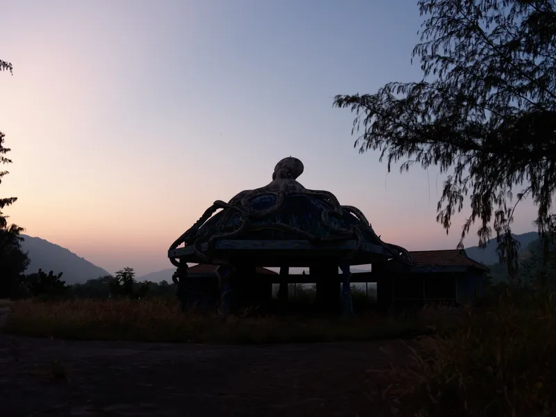 Silhouette of an octopus-shaped structure at dusk with soft pastel sky and surrounding trees, creating a mysterious and calm atmosphere in nature.