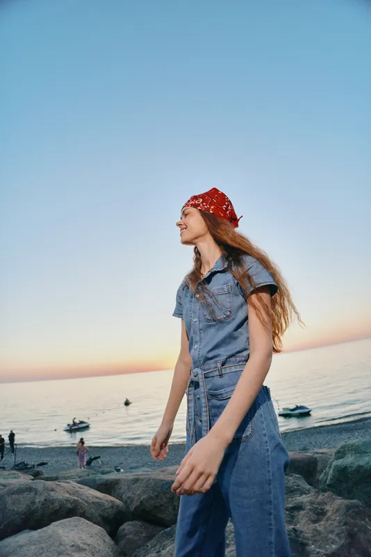 Lifestyle woman in modern boho-western denim street style with a red bandana, enjoying a serene beach sunset in soft film color tones and casual outfit.