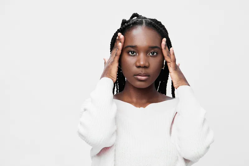 A thoughtful young Black woman in a cozy white sweater with braids, showing a pensive emotion against a neutral gray background