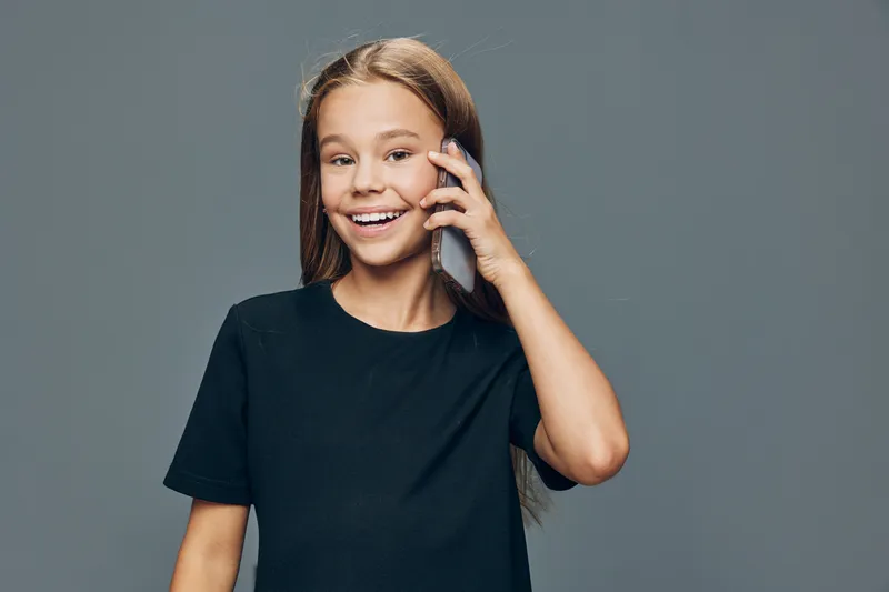 Girl with long hair smiling and talking on smartphone in casual black t shirt against grey background. Youth, communication and technology concept with happy child portrait.