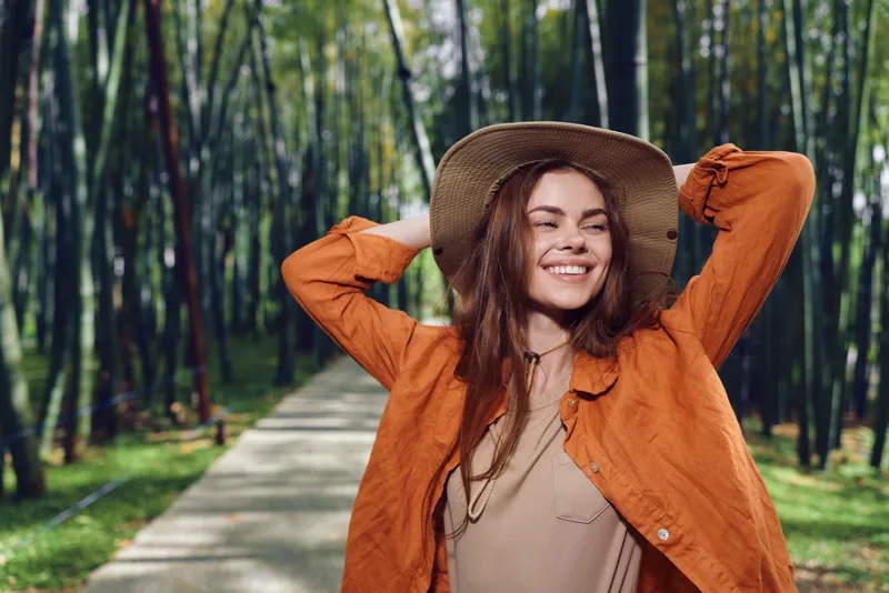 Woman smile hat jacket park path trees portrait outdoor nature: young female in orange jacket and straw hat smiling on a tree-lined park path, joyful portrait enjoying a sunny nature walk.
