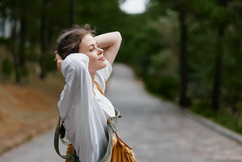 Young woman enjoying nature, wearing casual attire, smiling with eyes closed, surrounded by greenery in a serene outdoor setting