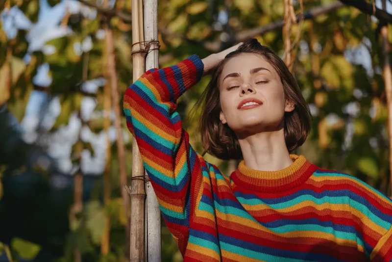 Woman portrait in rainbow sweater smiling with eyes closed outdoors in sunlight. Short hair model leans on wooden pole, relaxed expression, nature background.