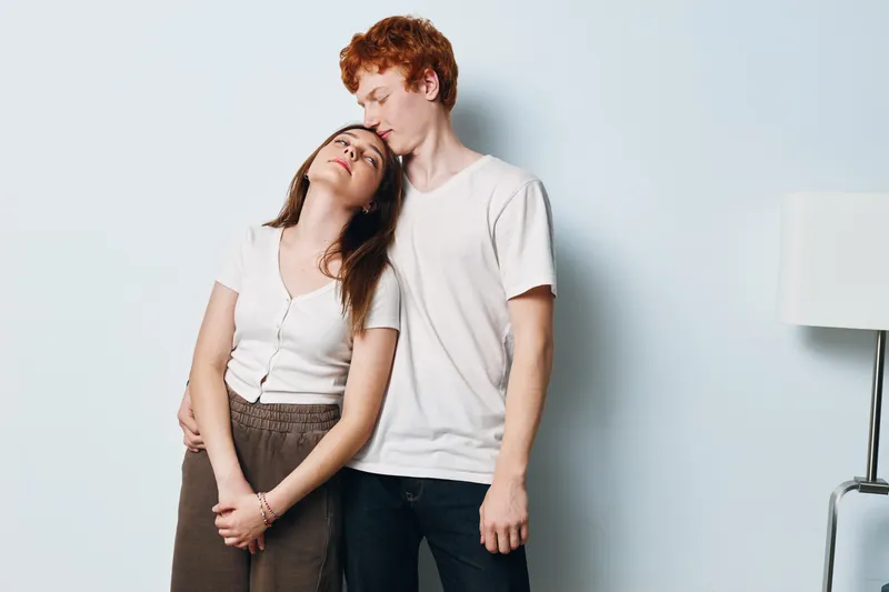 Couple in casual clothes hugging and showing affection indoors with white wall and lamp. Romantic young man and woman expressing love and tenderness in home environment.