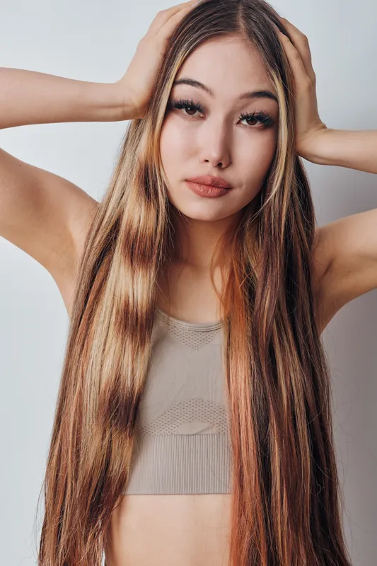 Young woman with long straight hair posing with hands on head wearing beige tank top with neutral expression on light background in studio portrait.