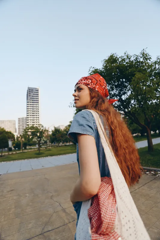 young woman with red hair wearing a bandana walks outdoors in an urban park surrounded by trees and modern buildings under a clear sky