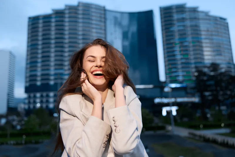 happy woman in beige coat laughing outdoors in urban business district with modern skyscrapers smiling with joy