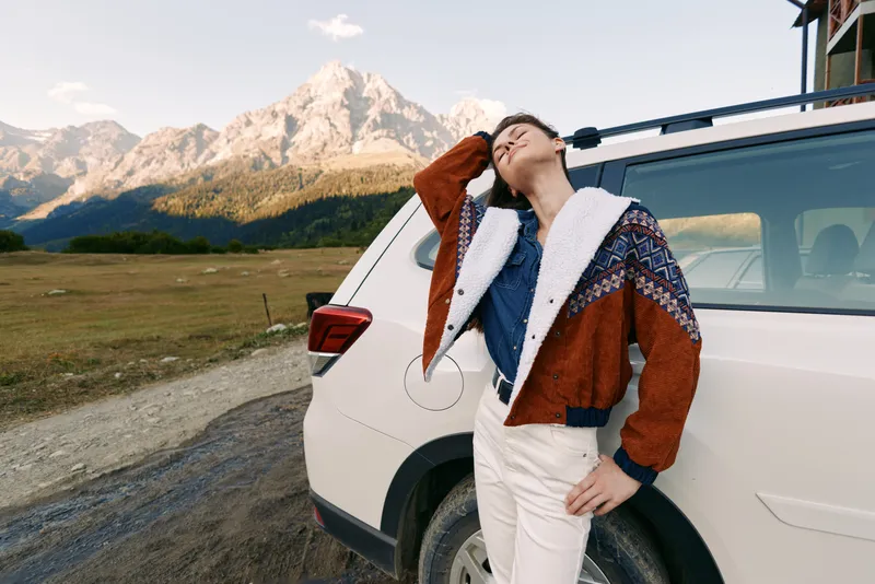 Woman by car on mountain road wearing warm jacket, looking up at peaks, travel and adventure scene in nature with SUV and scenic landscape for outdoor lifestyle portrait.