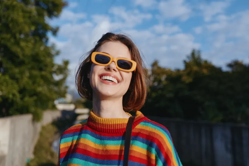Woman smiling in a rainbow sweater and yellow sunglasses, short hair, outdoor portrait on a sunny day. Joyful young adult in casual outfit, lifestyle travel vibe and confident energy.