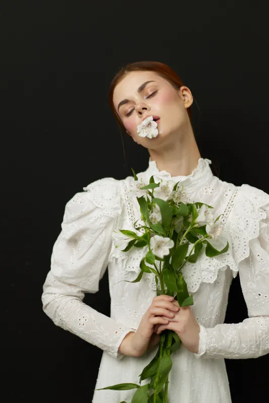 Elegant young woman in a white dress holding flowers against a black background, expressing tranquility and beauty with a serene expression