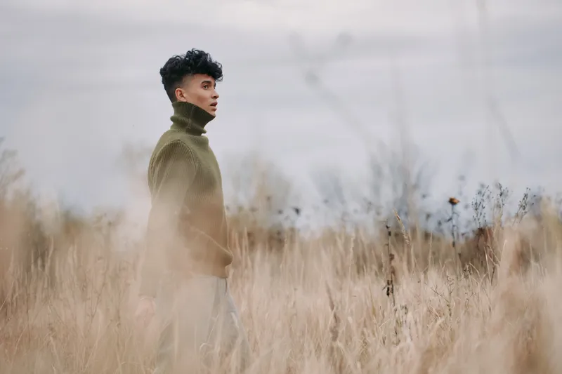 A thoughtful young man walks through tall dry grass in a knit turtleneck sweater, captured in soft natural light with muted tones to emphasize authenticity and quiet credibility in portrait.
